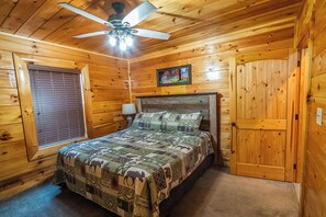 A wooden cabin bedroom with a bed, a ceiling fan, a window with blinds, and a door.