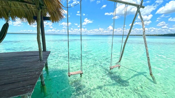 A wooden pier extending into clear turquoise water with a thatched roof and two swings.