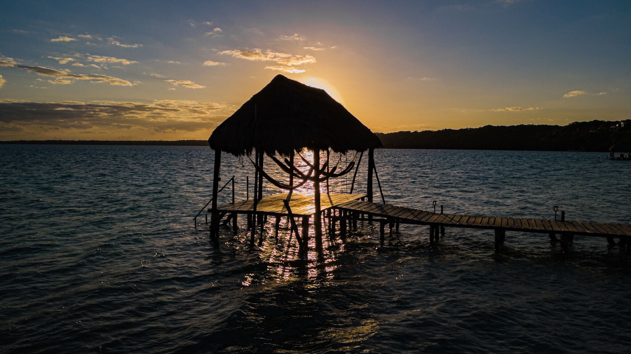 Un molo di legno che conduce a una capanna con tetto di paglia sopra l'acqua, con il sole che tramonta sullo sfondo.
