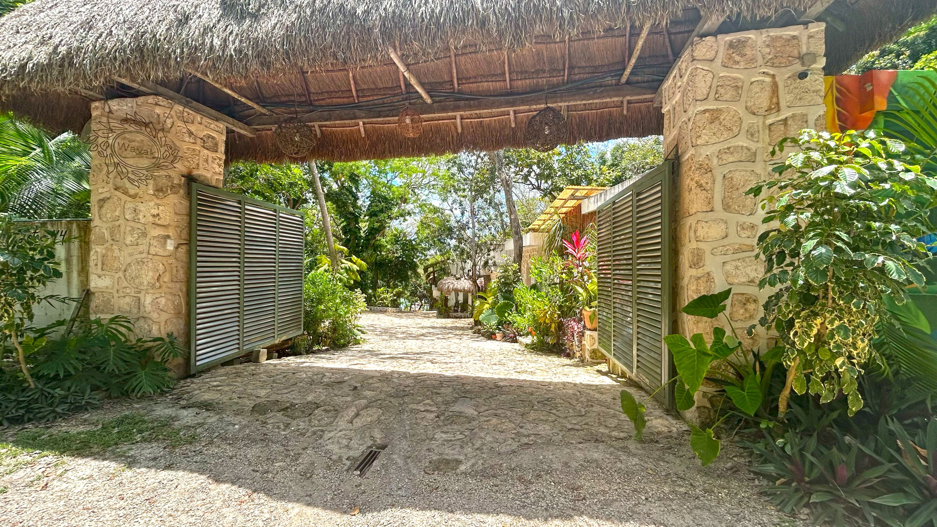 A stone archway leading to a covered area with a thatched roof, surrounded by lush greenery and potted plants.