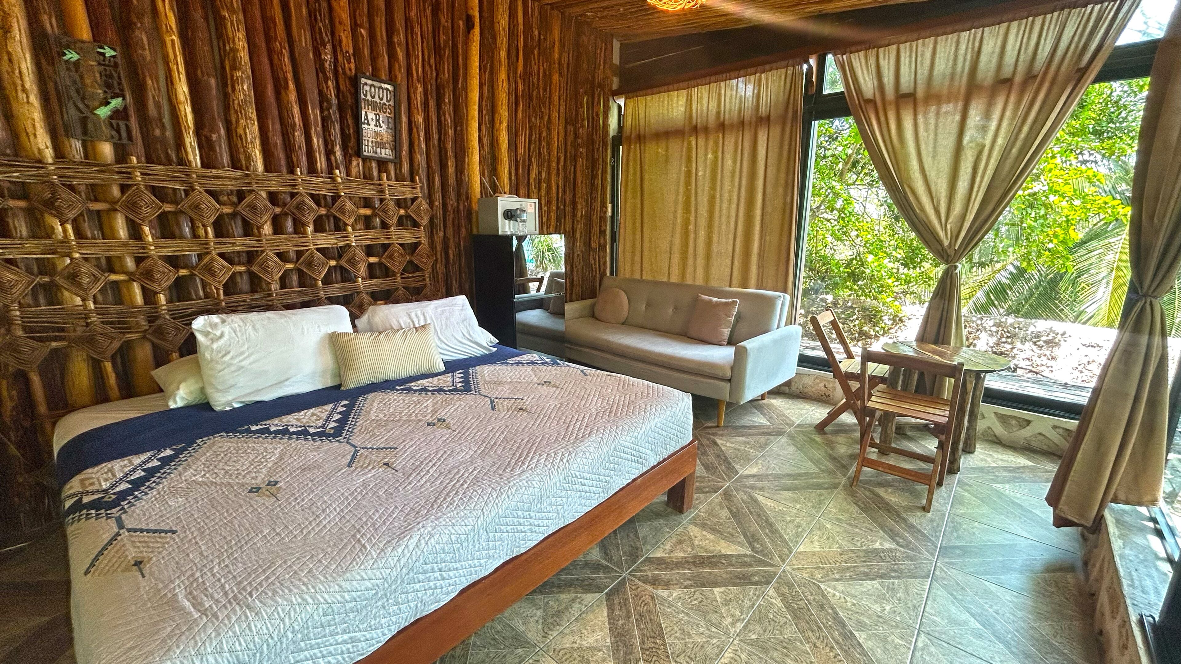 A room with a bed, a sofa, a small table, and a view of greenery outside.