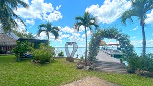 A wooden pier leading to a beach with palm trees, a thatched-roof structure, and a heart-shaped sculpture.