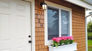A house with a white door, a window with a white window box containing pink flowers, and a brick exterior.