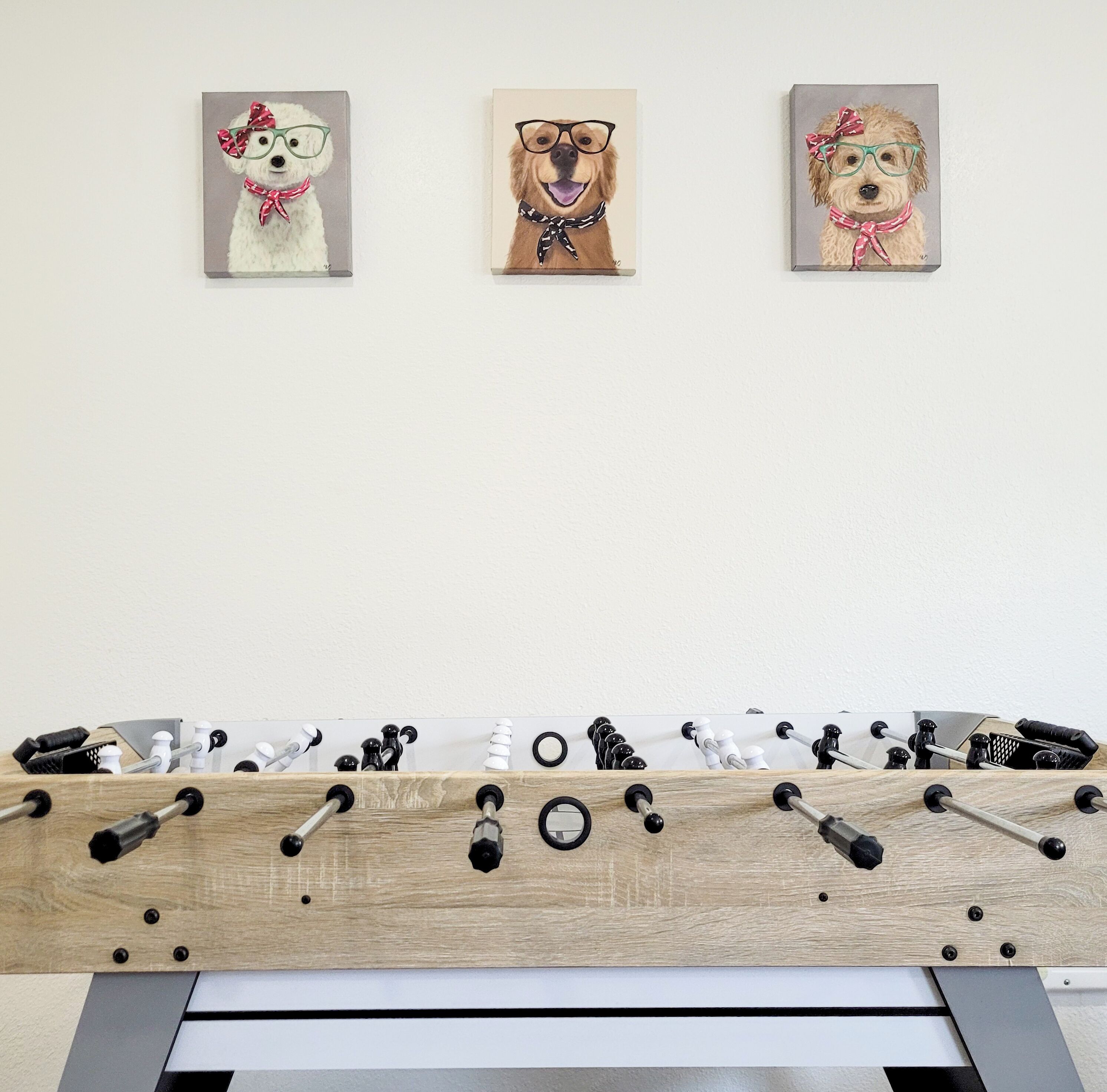 Une pièce dont le mur est décoré de trois tableaux représentant des chiens, et où l’on aperçoit un baby-foot au premier plan.