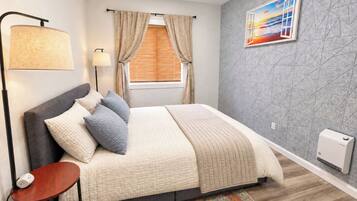 A modern bedroom with a bed, bedside lamp, window with curtains, and a painting on the wall.