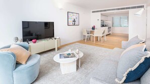 A modern living room with a flat-screen TV, a dining area, and a kitchen visible through an open doorway.