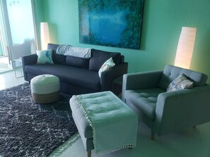 Ein Wohnzimmer mit einem dunkelgrauen Sofa, einem hellgrauen Sessel und passendem Hocker. Ein großes abstraktes Gemälde ziert die Wand, und im Hintergrund steht eine Stehlampe.