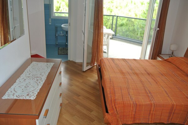 Une chambre avec une commode en bois, un lit recouvert d’une couverture orange, une salle de bain avec un lavabo et une chaise, et un balcon donnant sur un paysage verdoyant.