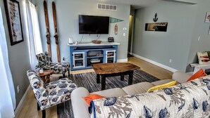 A living room with a sofa, coffee table, and TV. There is a fireplace, a ceiling fan, and a wall-mounted TV.