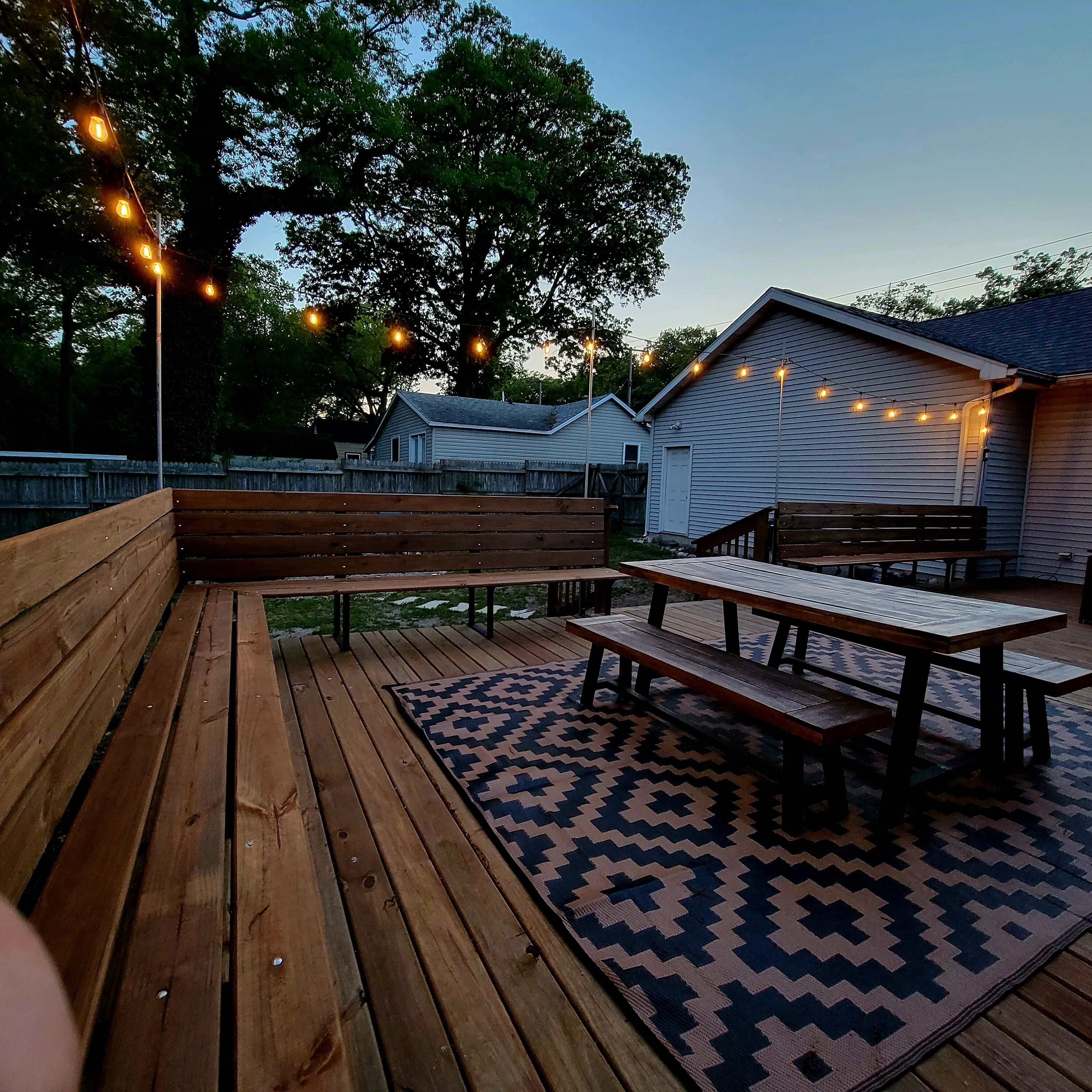 An outdoor patio area with wooden benches and picnic tables, string lights, and a patterned rug.
