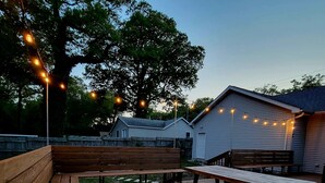 An outdoor patio area with wooden benches and picnic tables, string lights, and a patterned rug.
