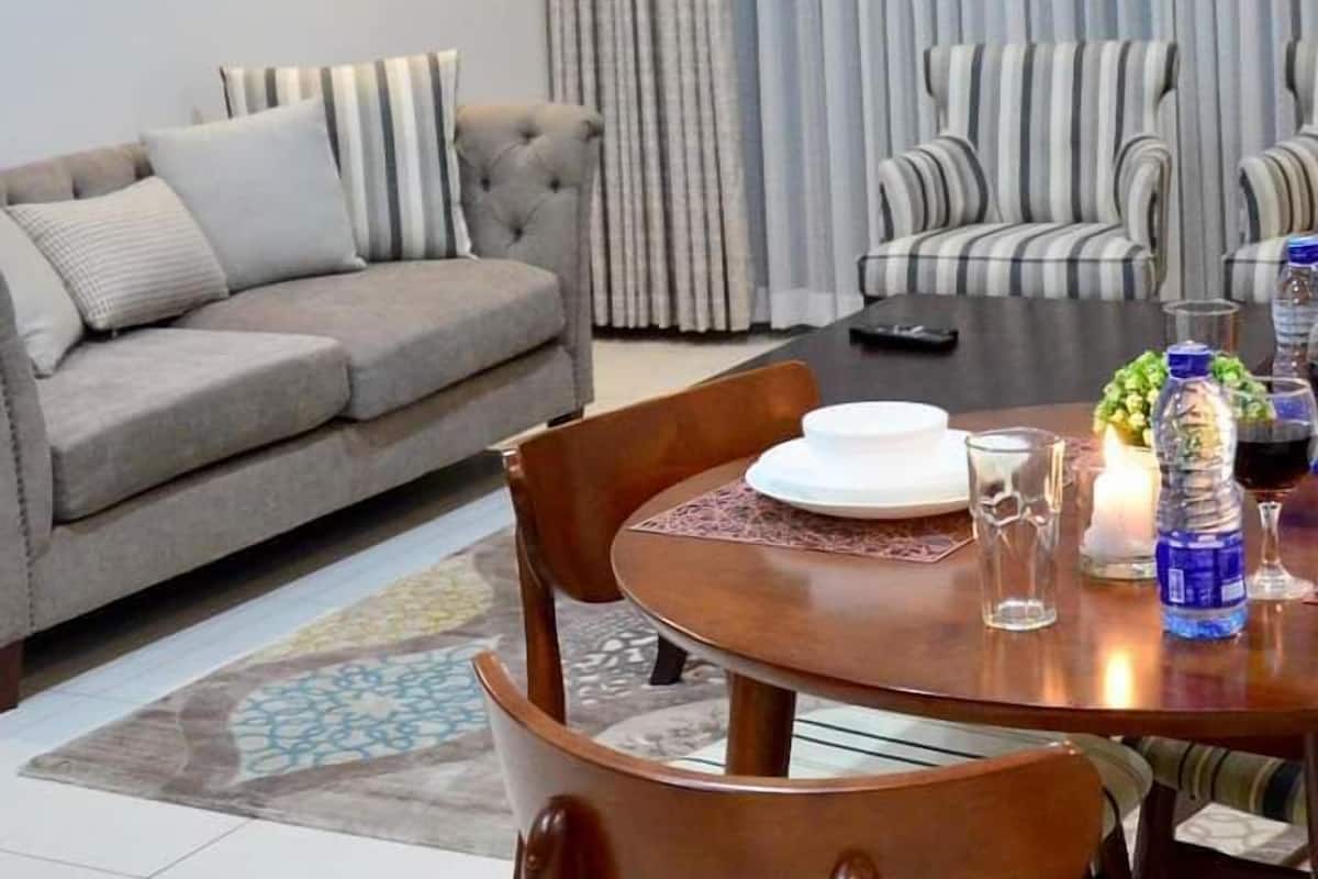 A modern living room with a grey sofa, striped armchairs, a wooden dining table with chairs, and a large window with sheer curtains.
