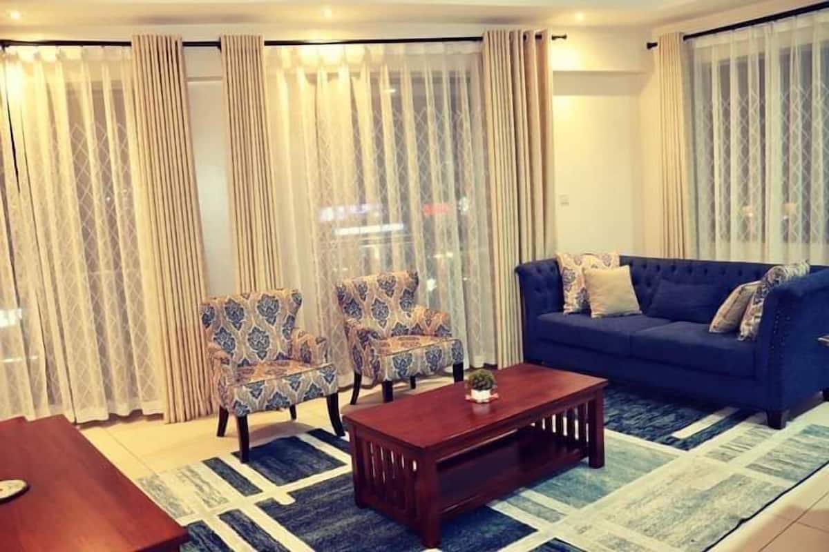 A living room with a blue sofa, patterned armchairs, a wooden coffee table, and a large window with sheer curtains.