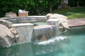 A swimming pool with a waterfall feature, surrounded by rocks and a basket of towels. - Walk 2 festivals Pool Spa Estate Sized Yard (Indio)