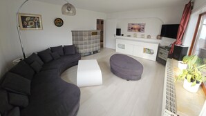 Ett modernt vardagsrum med en böjd svart soffa, en vit pall, en golvlampa, en köksdel med ett vitt skåp och en tv som är monterad på väggen.