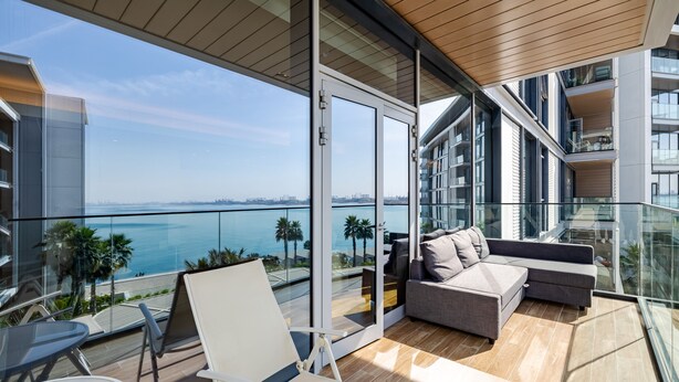 A modern balcony with a glass railing, a sofa, and a chair, overlooking a body of water and a cityscape. - DHH Bluewater Island Building 2 Sea View (Dubai)