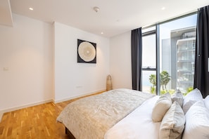 A modern bedroom with a large bed, a wall-mounted artwork, and a view of a building through a large window. - DHH Bluewater Island Building 2 Sea View (Dubai)