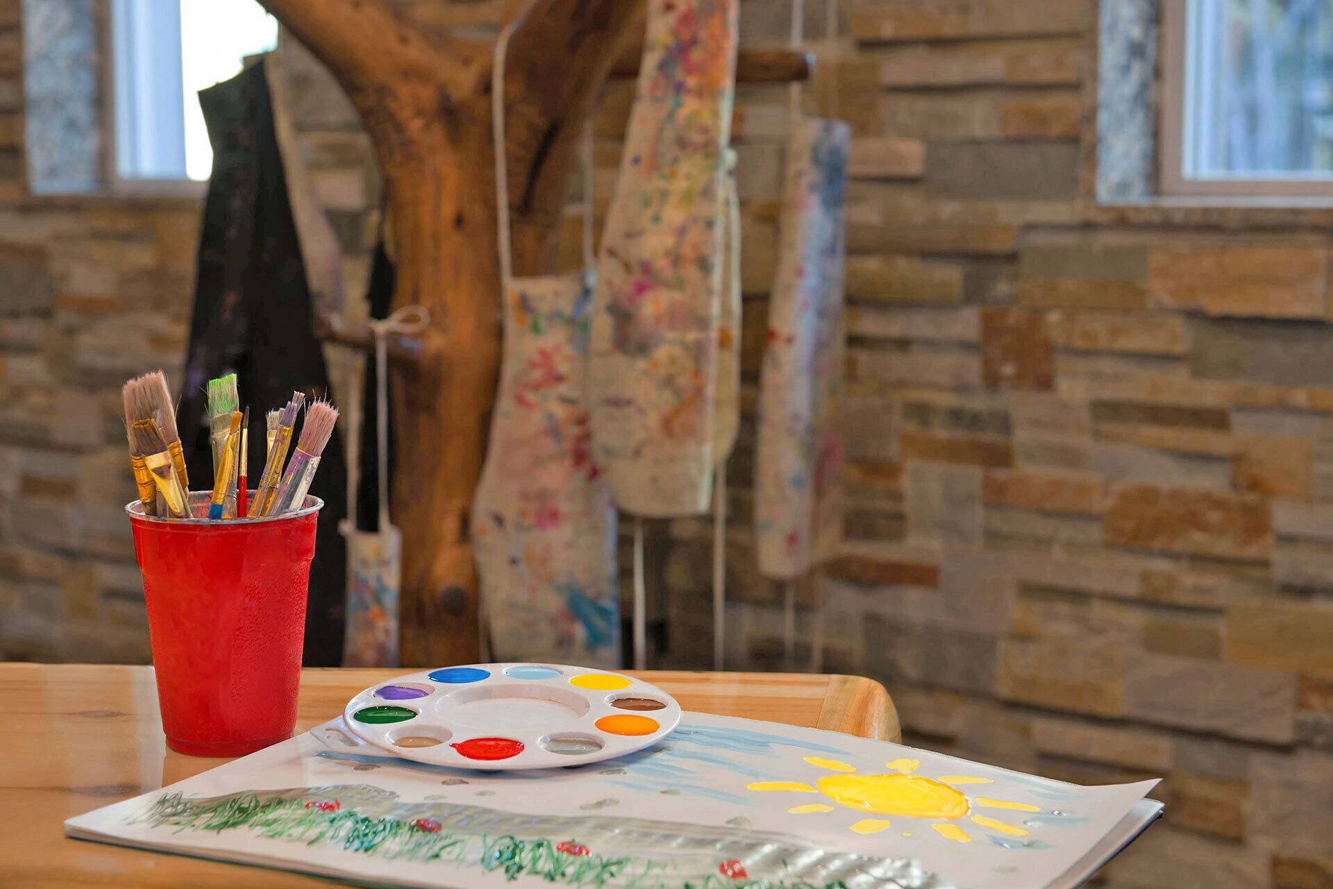 A painting of a sun on a table with a paint palette and brushes in a red cup.