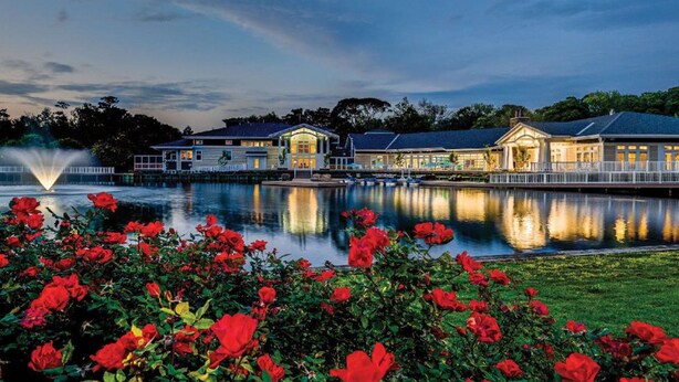A waterfront hotel with a reflective pool, a fountain, and lush red flowers in the foreground. - Packed with Amenities! Premier OBX Resort - Indoor Waterpark & Trails at Beachwoods Resort! (Kitty Hawk)
