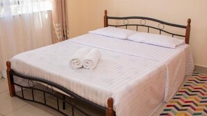 Una cama bien hecha con sábanas blancas y toallas enrolladas, un cabecero de madera y una alfombra colorida en el suelo.