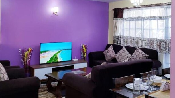 A living room with purple walls, a flat-screen TV, two sofas, a coffee table, and a dining area with a table and chairs. - Stay.Plus Karibu Homes Apartment (Athi River)