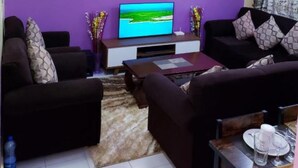 A living room with purple walls, a flat-screen TV, dark sofas, and a coffee table. - Stay.Plus Karibu Homes Apartment (Athi River)