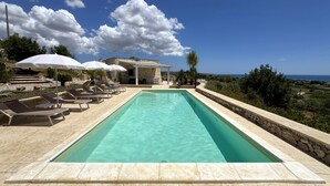 A rectangular pool with clear water, surrounded by lounge chairs and umbrellas, with a stone building and a view of the sea in the background.