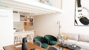 Ein modernes Wohnzimmer mit Esstisch aus Holz, grünen Sesseln, einer Couch und einem Couchtisch. Es gibt eine Küchenzeile mit weißen Schränken und einem hängenden Regal. An der Wand hängt ein großes abstraktes Gemälde.