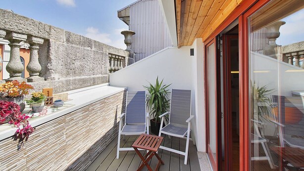 Ein Balkon mit Holzboden, weißen Wänden und einer Glastür zum Innenbereich. Zwei weiße Stühle und ein kleiner Tisch mit einem Blumentopf stehen bereit.