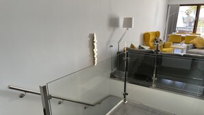 Uma escada moderna com guarda-corpos de vidro e corrimão metálico, que dá acesso a uma sala de estar com sofá e poltronas amarelas.