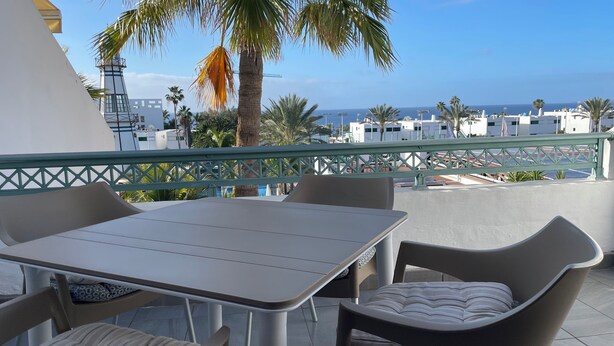 Terraço com mesa e cadeiras, com vista para uma área costeira com edifícios e palmeiras.