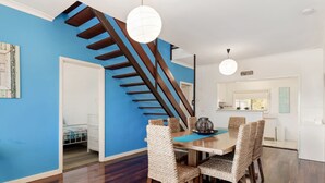 A dining area with a wooden table and woven chairs, a staircase, and a kitchen in the background. - Giggles Beach House in Silver Sands (Silver Sands)