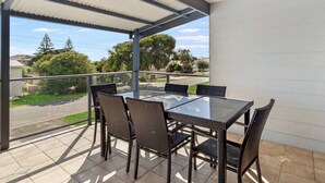 A covered outdoor patio with a glass table and wicker chairs, overlooking a residential street. - Giggles Beach House in Silver Sands (Silver Sands)