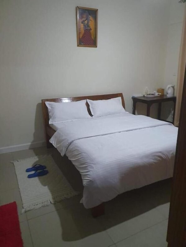 Une chambre à coucher avec un lit, une petite table sur laquelle se trouve une bouilloire, et un tableau accroché au mur.