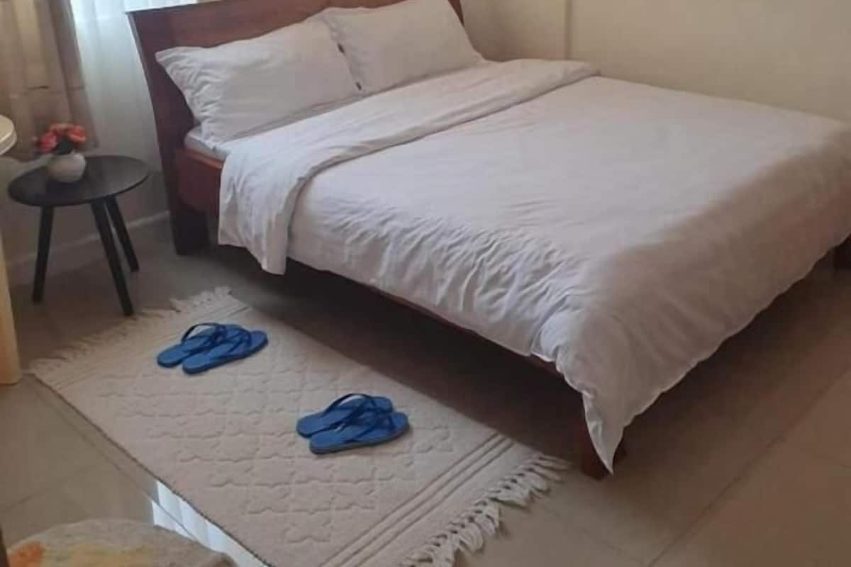 A bedroom with a bed, a small round table with a vase, and a pair of blue slippers on a rug.