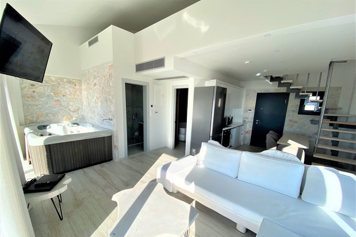 A modern hotel room with a large white sofa, a small table, a bathtub, and a TV mounted on the wall.