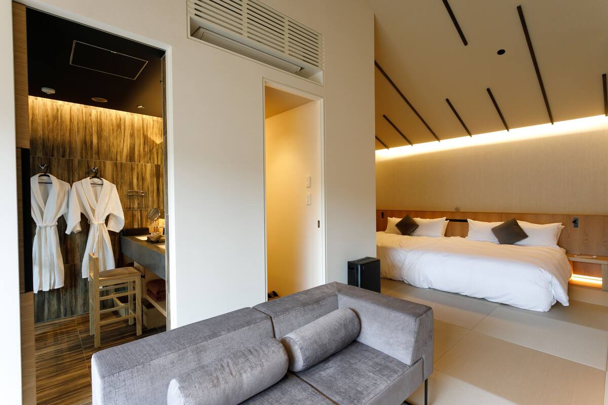 Una habitación de hotel moderna con una cama grande, un sofá gris, un vestidor con batas y suelo de madera.