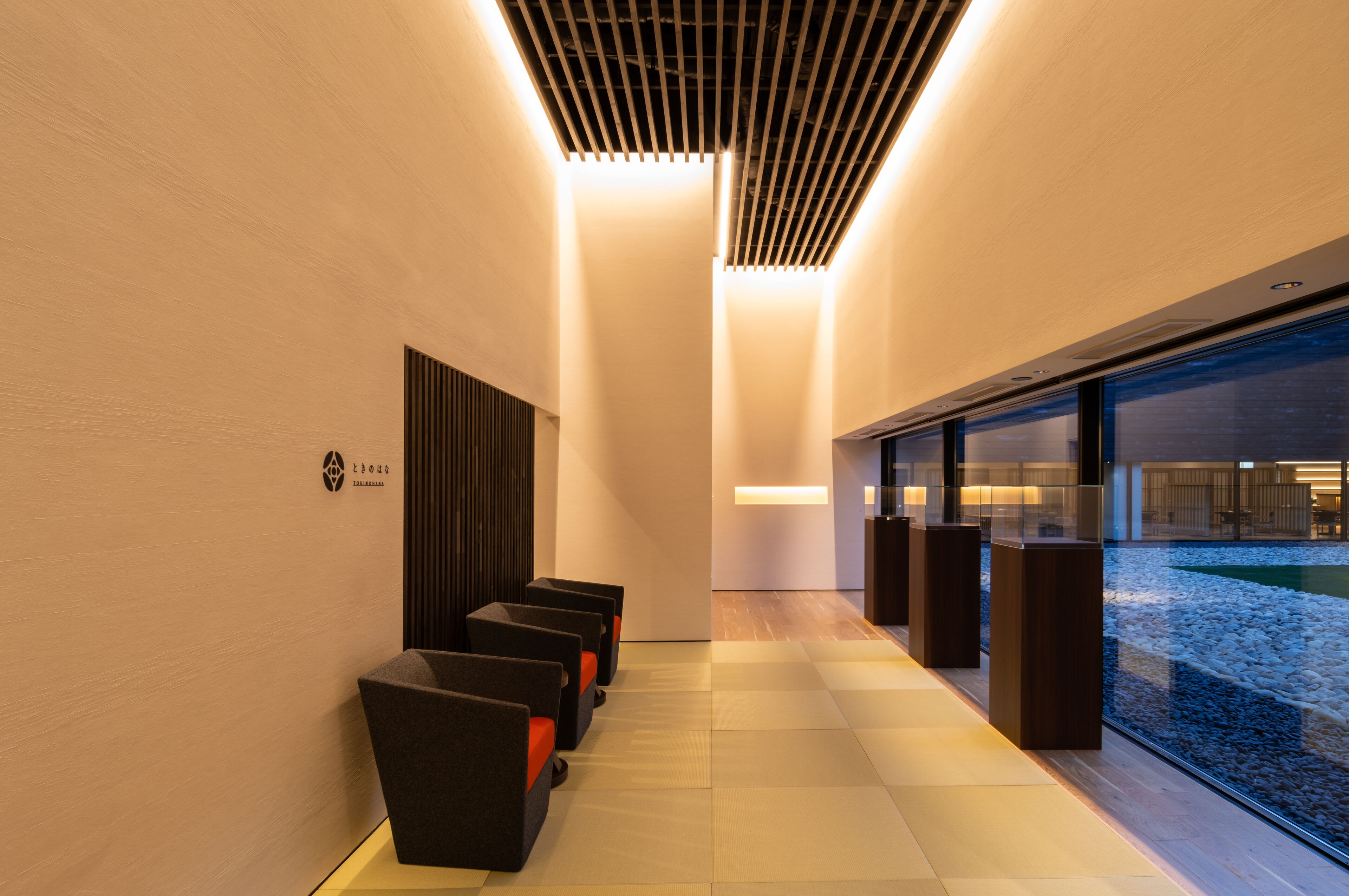 A modern hallway with a row of black chairs, a wooden slatted ceiling, and a glass wall with a view of a water feature.