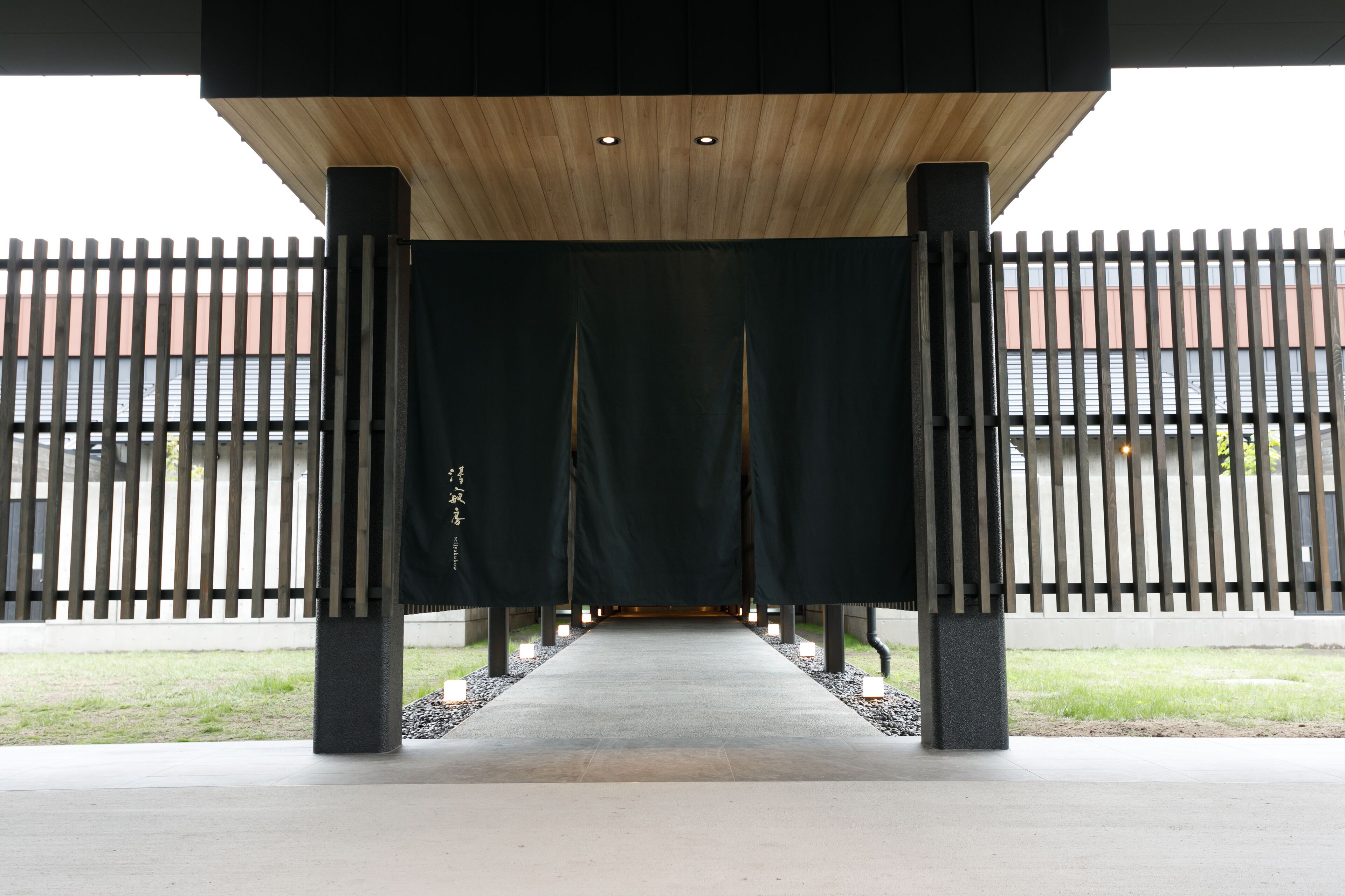 A covered walkway with a wooden ceiling and black curtains, leading to a building with a fence.
