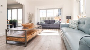 Un salon spacieux avec un canapé gris, une table basse en bois et un divan gris. Une grande fenêtre laisse entrer la lumière naturelle et offre une vue sur l’extérieur.
