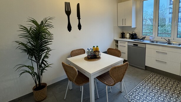 A modern kitchen with white cabinets, a dining table with four wicker chairs, a potted plant, and wall-mounted utensils. - Pleasant townhouse between Paris and Disneyland park (Vaires-sur-Marne)