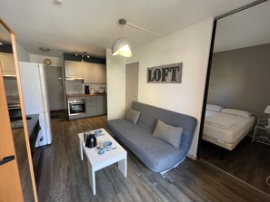 Ein kompakter Wohnraum mit Küchenzeile, einem grauen Sofa, einem kleinen weißen Tisch und einem Bett.