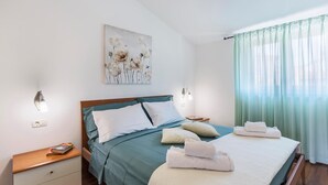Une chambre à coucher bien rangée, avec un lit, des tables de chevet, une lampe fixée au mur, une prise électrique et une fenêtre ornée de rideaux légers.