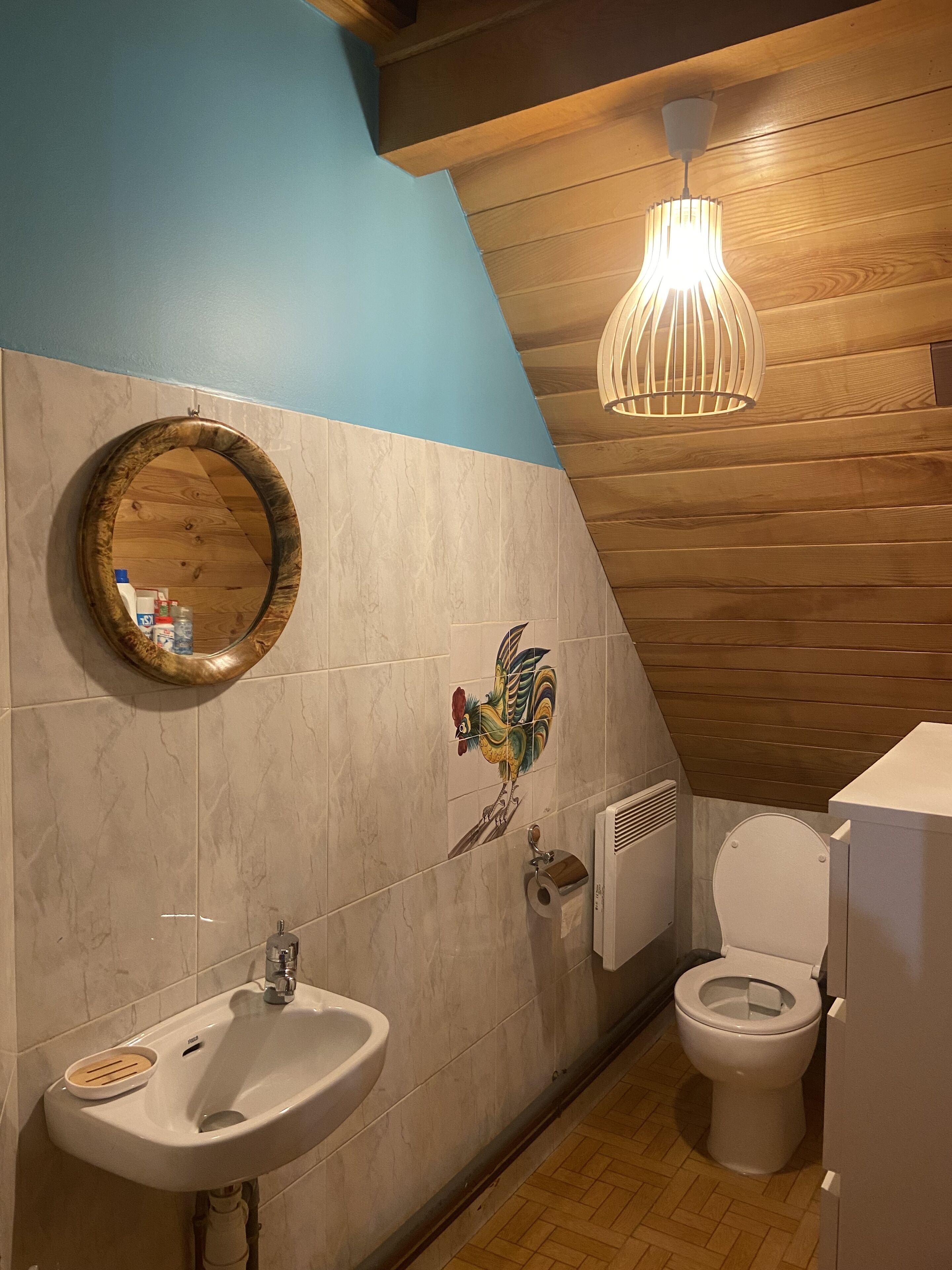 Un baño con techo de madera, un lavabo blanco, un inodoro, un espejo con marco de madera y un azulejo decorativo con forma de gallo.