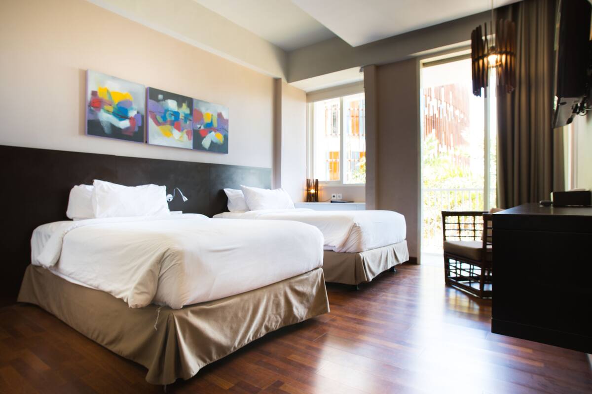 A hotel room with two beds, a desk, and artwork on the wall.