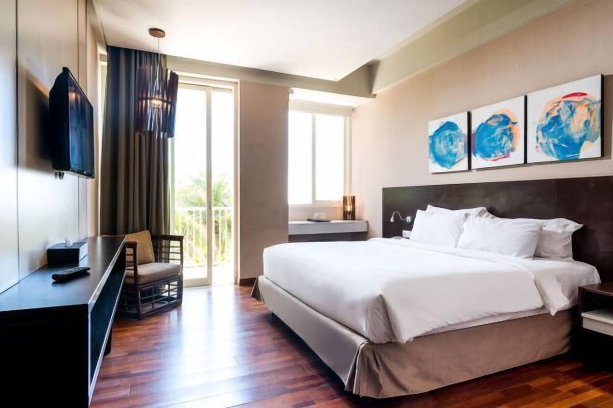 A modern hotel room with a large bed, a desk, a TV, and artwork on the wall.