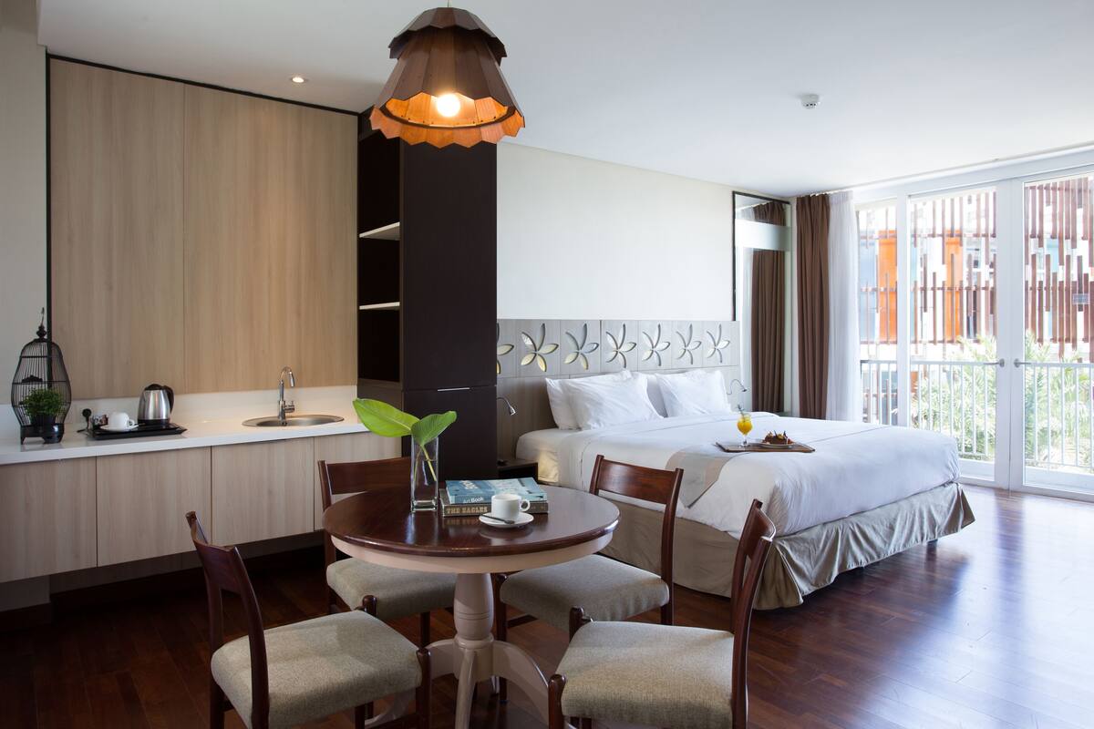 A hotel room with a bed, a dining table with chairs, a kitchen area, and a large window with a view.
