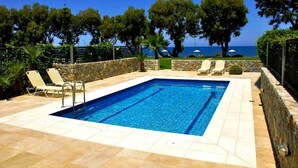Ein rechteckiger Swimmingpool mit einer Steinmauer und einer gefliesten Liegefläche, umgeben von Liegestühlen und Blick auf das Meer.