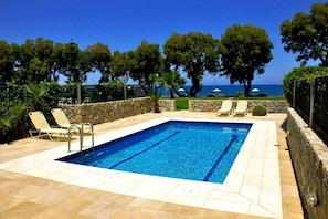A rectangular swimming pool with a stone wall and tiled deck, surrounded by lounge chairs and a view of the sea.