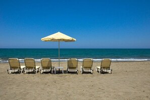 A row of beach chairs and a single umbrella on a sandy beach with a clear blue sky and ocean in the background.